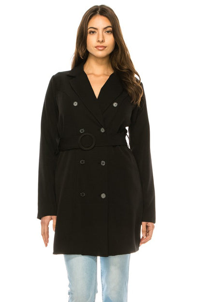 A woman wearing a long black coat with big buttons, a white top, and blue jeans.  Background's plain white with a black border.