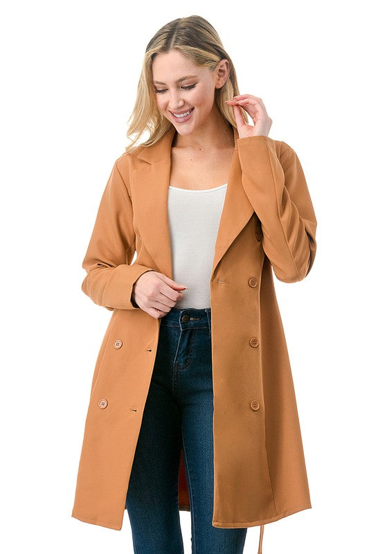 A woman wearing a long tan coat with big buttons, a white top, and blue jeans.  Background's plain white with a black border.
