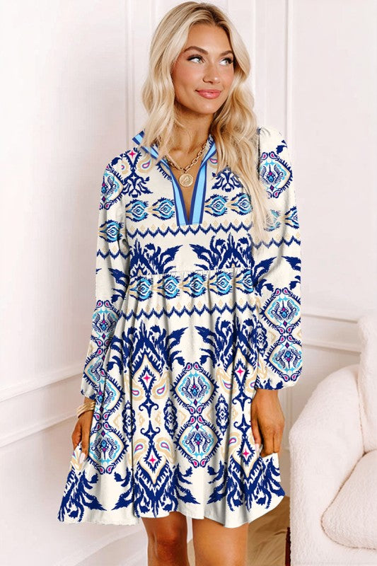 Woman wearing a blue ethnic print mini dress with striped patterns, split neckline, and long sleeves, standing indoors against a white paneled wall.
