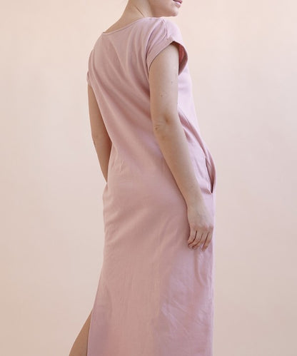 A woman wearing a light pink, short-sleeve, midi-length casual dress with side pockets, paired with black flat sandals.