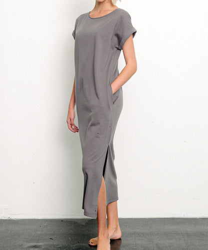 Woman in a stylish gray dress with a side slit, posing against a minimalist white backdrop. One hand is tucked into her pocket, while the other hangs relaxed by her side, with bare feet adding a touch of casual elegance.