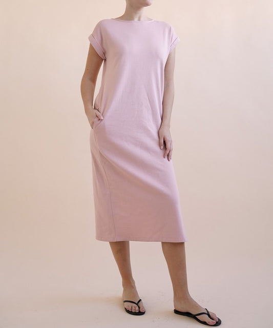 A woman wearing a light pink, short-sleeve, midi-length casual dress with side pockets, paired with black flat sandals.