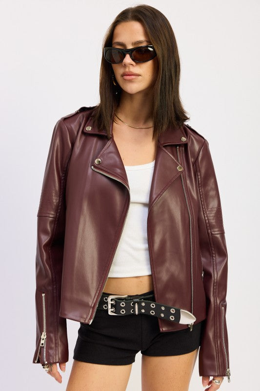 Woman in burgundy leather jacket with white top and black shorts against beige background