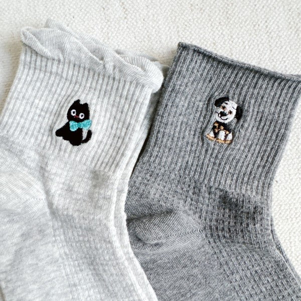 A pair of crew socks in light grey and dark grey. The light grey sock features a small embroidered black cat with a blue bow, while the dark grey sock has an embroidered dog design.