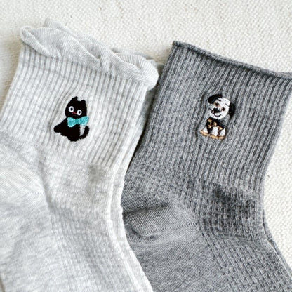 A pair of crew socks in light grey and dark grey. The light grey sock features a small embroidered black cat with a blue bow, while the dark grey sock has an embroidered dog design.