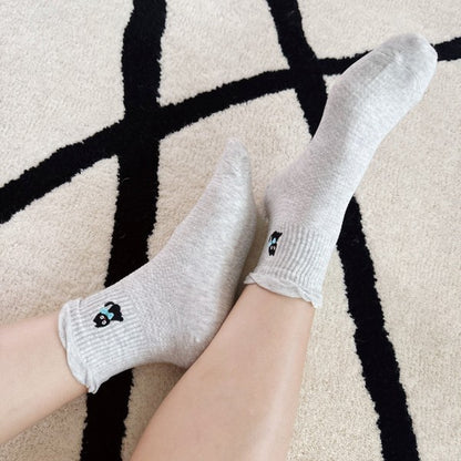 An image showing someone wearing a pair of crew socks in light grey, It features a small embroidered black cat with a blue bow design. 