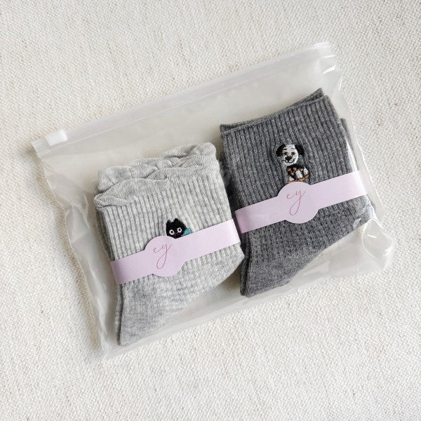 A pair of crew socks in light grey and dark grey neatly packed. The light grey sock features a small embroidered black cat with a blue bow, while the dark grey sock has an embroidered dog design.