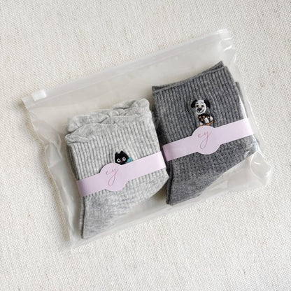 A pair of crew socks in light grey and dark grey neatly packed. The light grey sock features a small embroidered black cat with a blue bow, while the dark grey sock has an embroidered dog design.