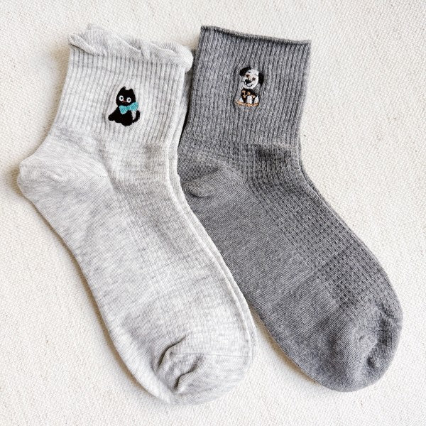 A pair of crew socks in light grey and dark grey. The light grey sock features a small embroidered black cat with a blue bow, while the dark grey sock has an embroidered dog design.