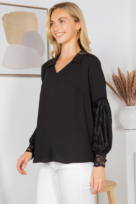 A woman with long blonde hair is posing in a black blouse with frilly sleeves and white pants. She's sitting on a wooden stool with one hand on her hip. There's a white wall behind her with a framed picture and some dried pampas grass in a vase.