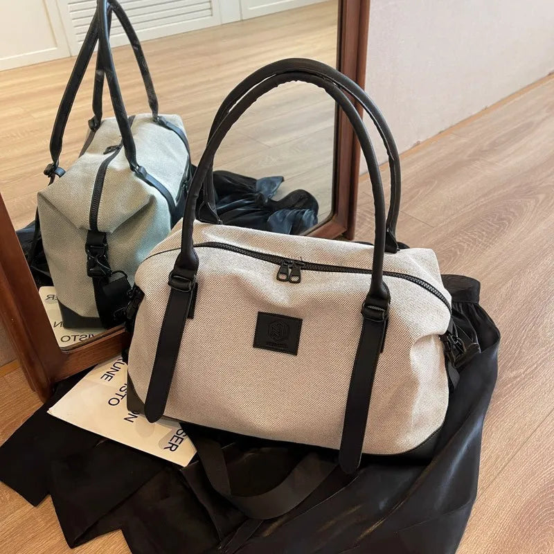 Light-colored handbag with black handles, accents, and front logo patch—placed on a wooden floor in front of a mirror, with a black cloth and white paper beneath it.