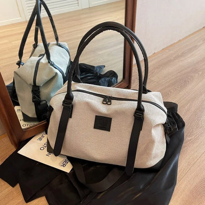 Light-colored handbag with black handles, accents, and front logo patch—placed on a wooden floor in front of a mirror, with a black cloth and white paper beneath it.