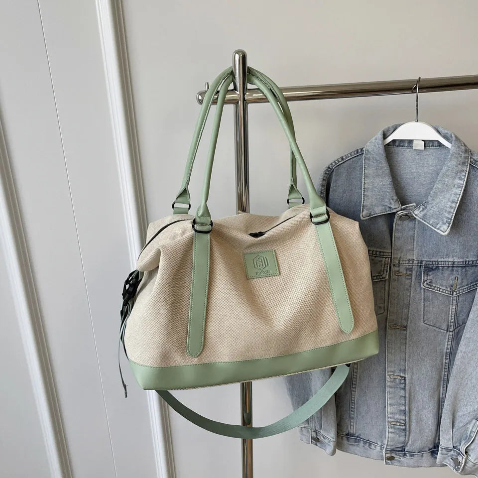 Beige and green handbag with green handles, base, and front logo patch—hanging beside a denim jacket on a metal rack in a clean indoor setting.