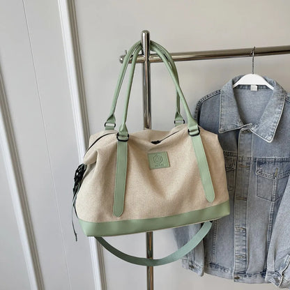 Beige and green handbag with green handles, base, and front logo patch—hanging beside a denim jacket on a metal rack in a clean indoor setting.