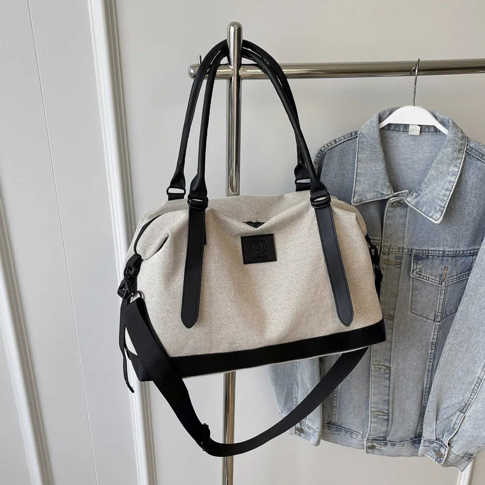 Beige and black duffel bag with black leather handles, shoulder strap, and front logo patch—hanging beside a denim jacket on a metal rack in a minimalist indoor setting.