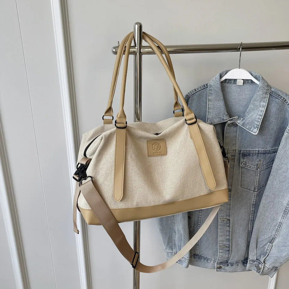 Beige and tan handbag with top handles, detachable shoulder strap, and front logo patch—hanging beside a denim jacket on a metal rack in a clean indoor setting.