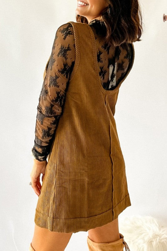 Side view of a woman wearing a brown corduroy sleeveless mini dress with front patch pockets, styled over a black lace long-sleeve top and paired with tan knee-high boots