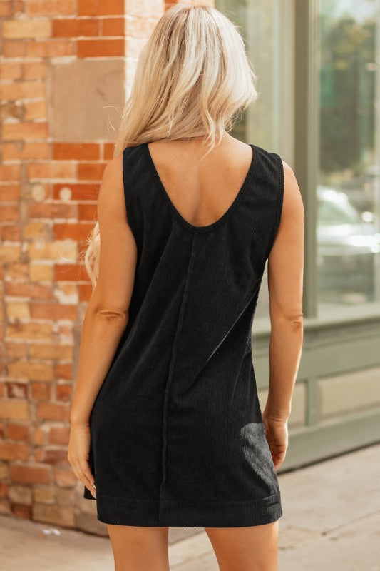 A black corduroy sleeveless mini dress with front patch pockets, styled over a black lace long-sleeve top and paired with tan knee-high boots.
