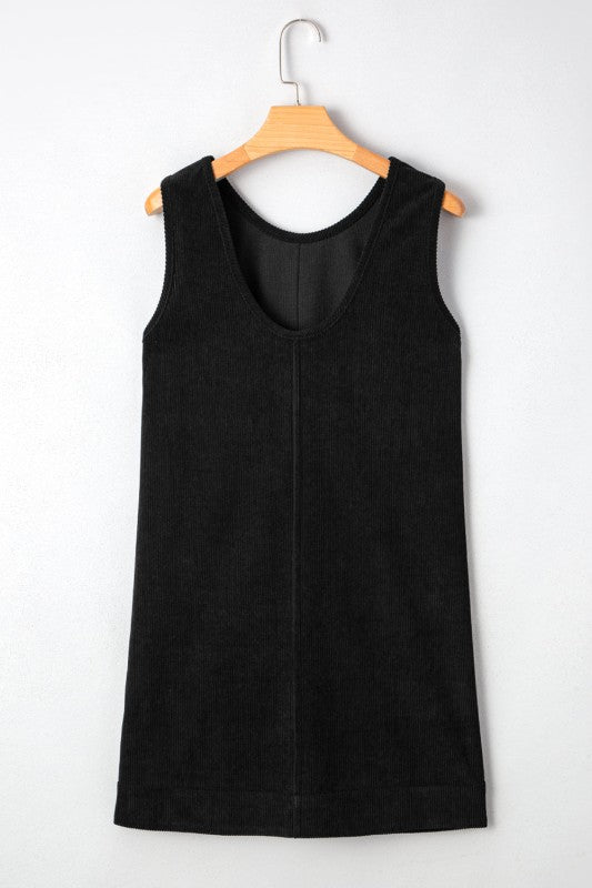 A black corduroy sleeveless mini dress with front patch pockets, styled over a black lace long-sleeve top and paired with tan knee-high boots.
