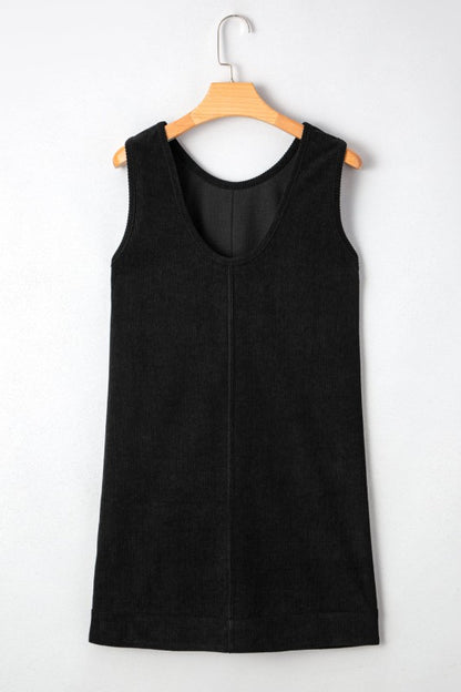 A black corduroy sleeveless mini dress with front patch pockets, styled over a black lace long-sleeve top and paired with tan knee-high boots.
