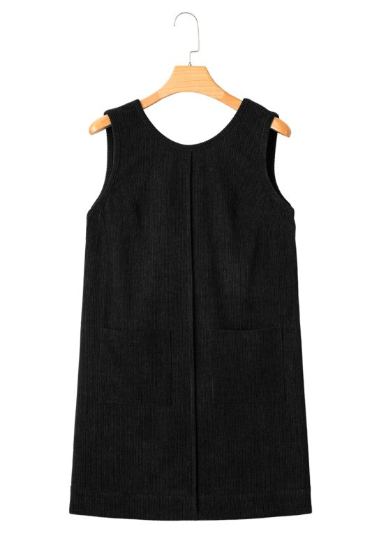 A black corduroy sleeveless mini dress with front patch pockets, styled over a black lace long-sleeve top and paired with tan knee-high boots.
