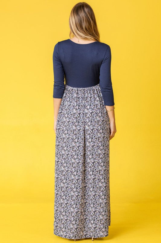 A woman wearing a navy blue top with a floral maxi skirt, posing in a yellow background.