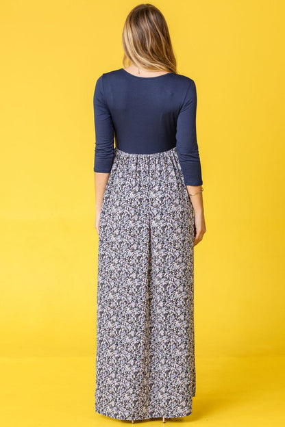 A woman wearing a navy blue top with a floral maxi skirt, posing in a yellow background.