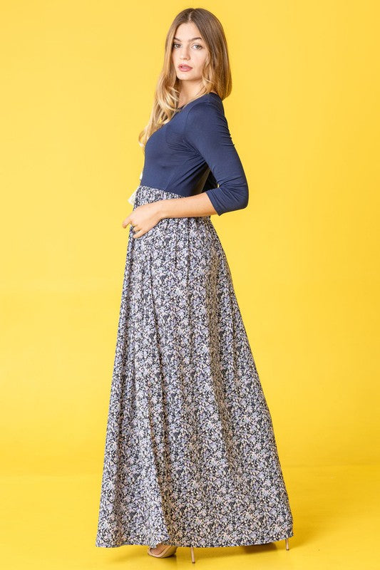 A woman wearing a navy blue top with a floral maxi skirt, posing in a yellow background.