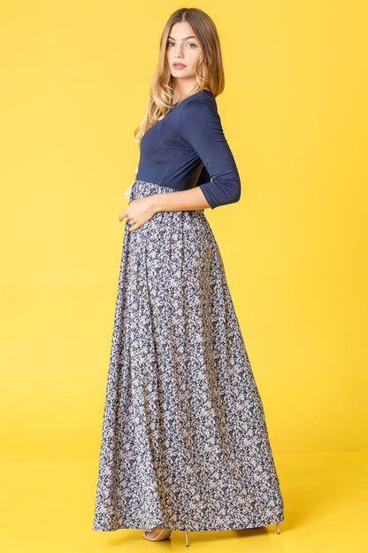A woman wearing a navy blue top with a floral maxi skirt, posing in a yellow background.