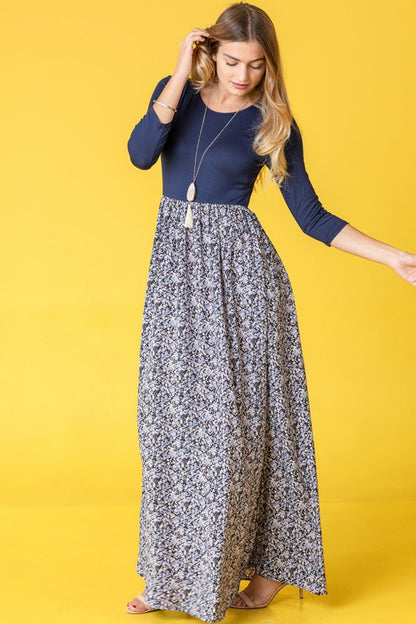 A woman wearing a navy blue top with a floral maxi skirt, posing in a yellow background.