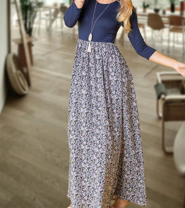 A woman wearing a navy blue top with a floral maxi skirt is posing in a room with wooden floors and dining furniture.