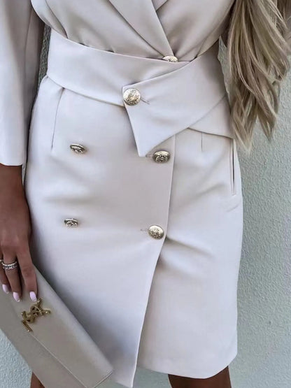 A woman in a stylish white double-breasted blazer dress with gold buttons, accessorized with jewelry and a clutch. 