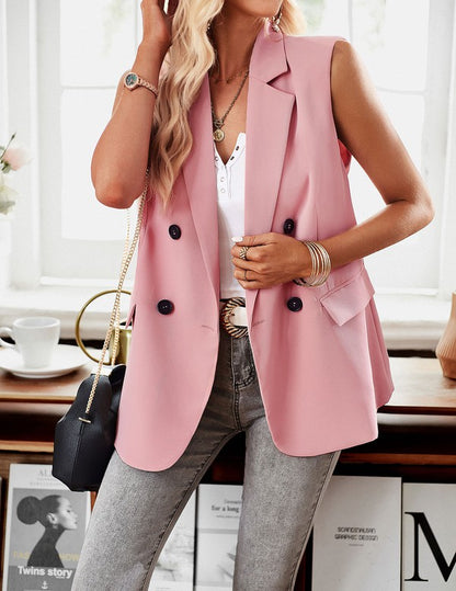 A woman posing in a stylish outfit. She's wearing a pink sleeveless blazer over a white top, paired with light gray jeans and accessorized with gold jewelry and a black purse.