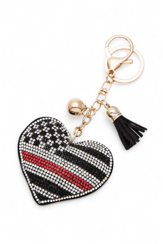 This keychain features a heart-shaped pendant with a design resembling the American flag but with a twist - a thin red line through it. The heart is made of rhinestones in black, white, and red. It has a gold chain with a black tassel and a gold keyring attachment.