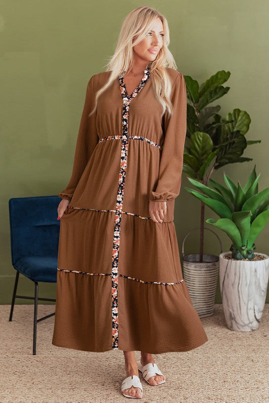 Woman wearing a long brown tiered maxi dress with floral trim accents, long sleeves, and a relaxed fit, styled with white sandals.”