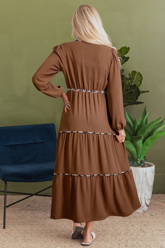 Back view of a woman wearing a long brown tiered maxi dress with floral trim accents, long sleeves, and a relaxed fit, styled with white sandals.