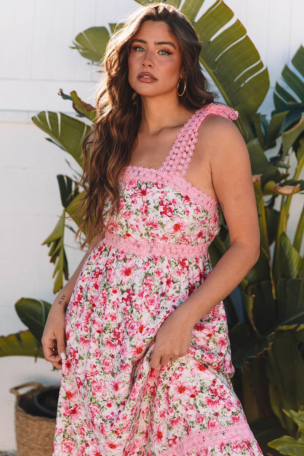 Woman wearing a pink floral maxi dress with lace-trimmed straps, posing in front of tropical plants on gravel.