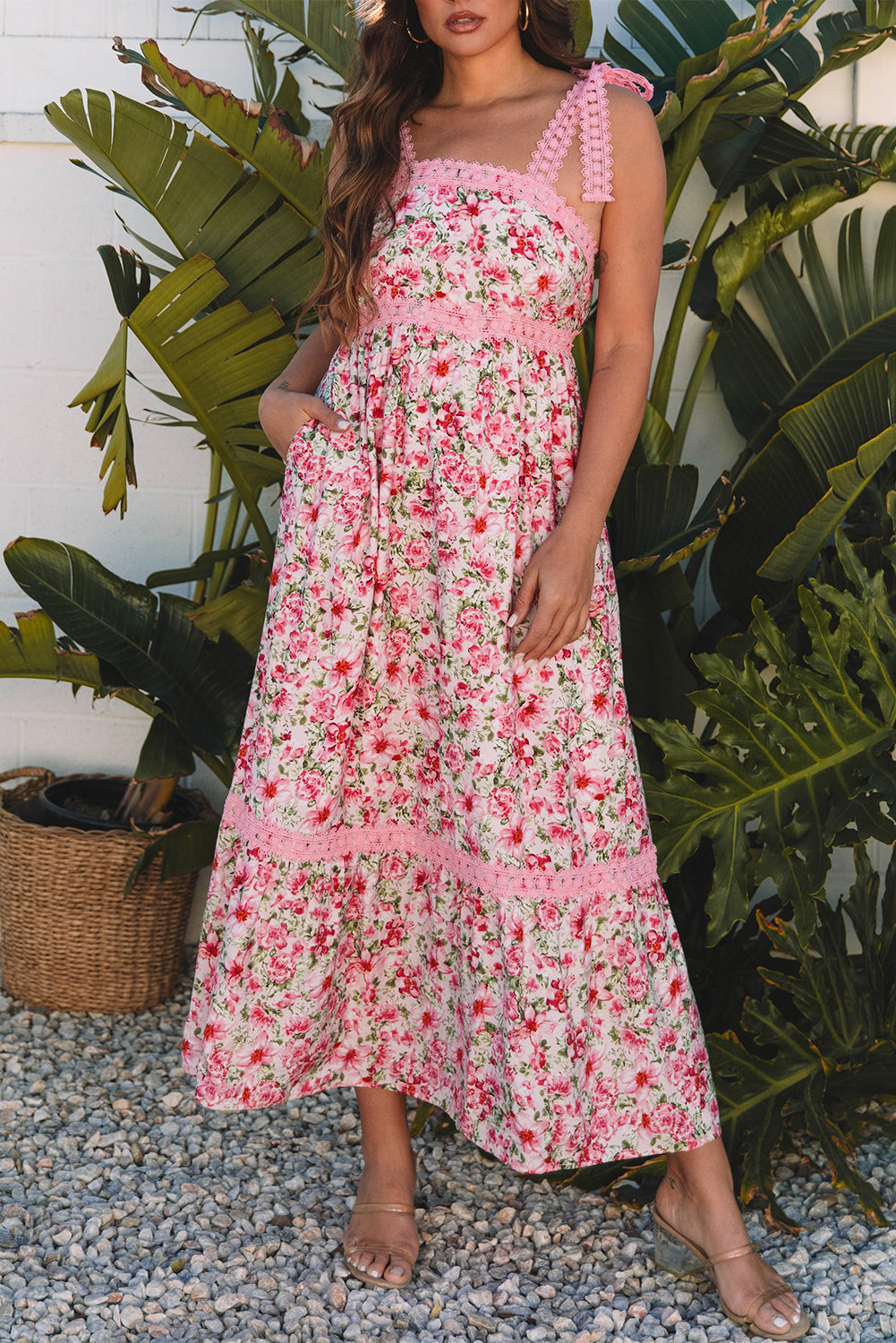 Woman wearing a pink floral maxi dress with lace-trimmed straps, posing in front of tropical plants on gravel.