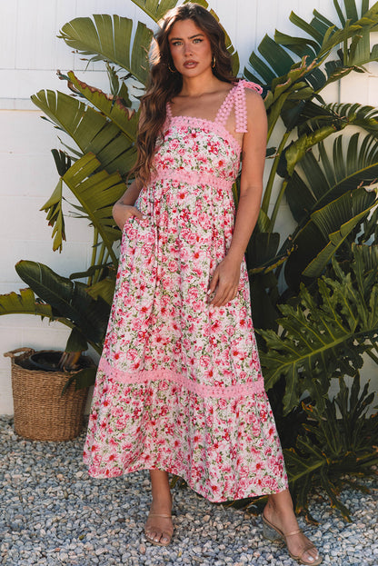 Woman wearing a pink floral maxi dress with lace-trimmed straps, posing in front of tropical plants on gravel.
