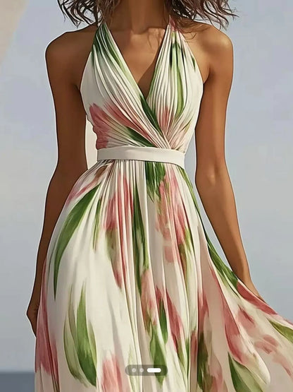 A woman wearing a flowy, white maxi dress with pink flowers and green leaves. It's a halter neck dress with a V-neckline and a belt at the waist. She's standing on a beach with the ocean behind her.