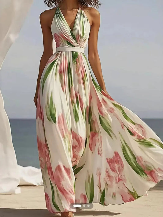 A woman wearing a flowy, white maxi dress with pink flowers and green leaves. It's a halter neck dress with a V-neckline and a belt at the waist. She's standing on a beach with the ocean behind her.