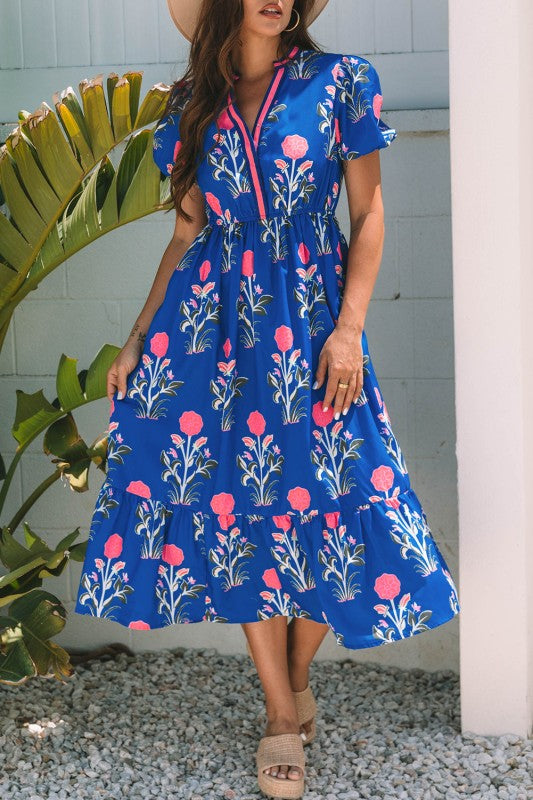 A woman posing in a vibrant blue dress with pink flowers and green leaves printed on it. She's wearing a hat, hoop earrings, and tan heels, standing on gravel next to a plant and a white wall.