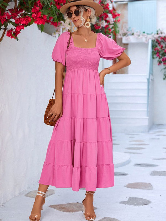 A woman in a vibrant pink maxi dress with puffy sleeves and a square neckline poses in a picturesque setting. She's wearing a tan hat, sunglasses, and strappy sandals, holding a brown purse. 