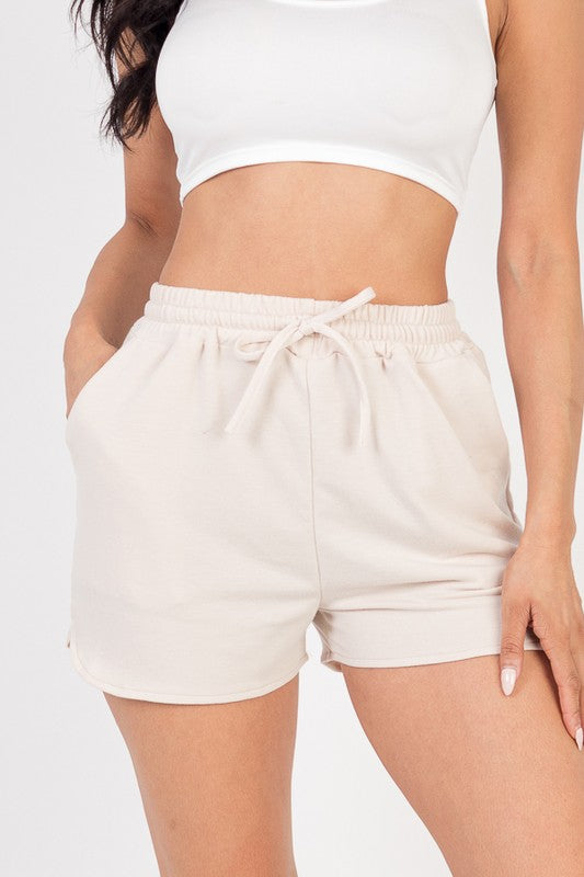 The image shows a woman wearing a white crop top and off-white shorts. She's posing with one hand in her pocket and the other on her thigh, against a plain white background.