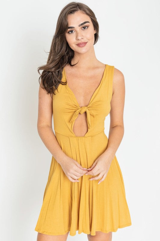 Woman in a mustard yellow sleeveless mini dress with a front knot, deep V-neckline, and cut-out detail below the bust.
