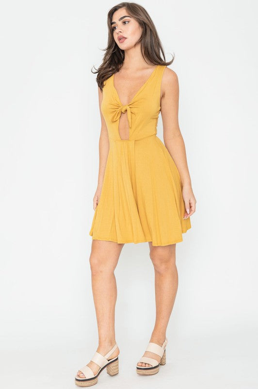Woman in a mustard yellow sleeveless mini dress with a front knot, deep V-neckline, and cut-out detail below the bust.
