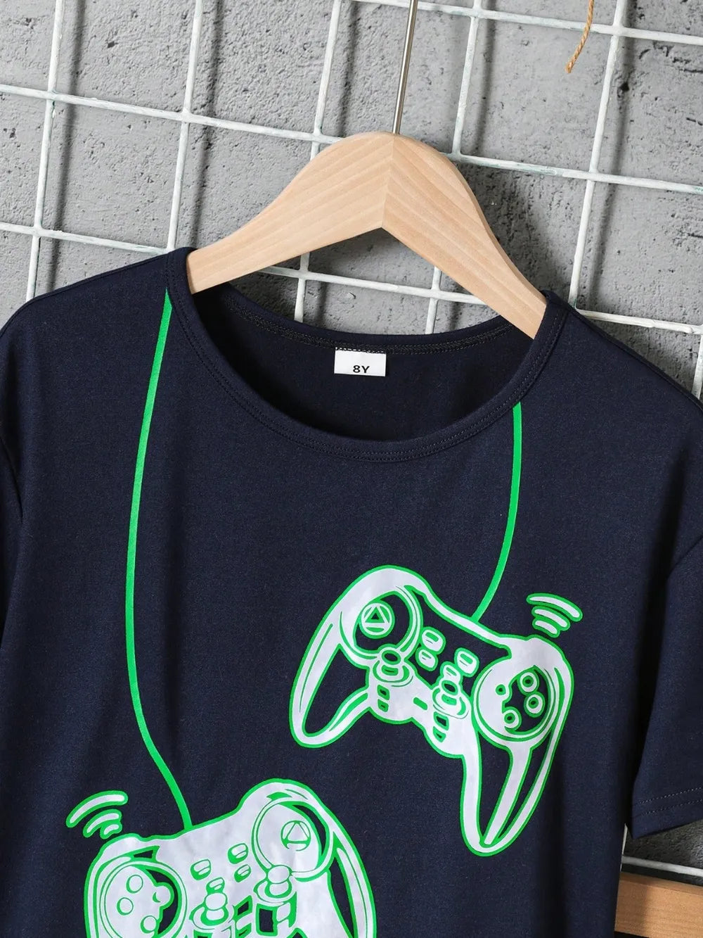 Navy blue t-shirt with green gaming controller design on a hanger against a grid wall.
