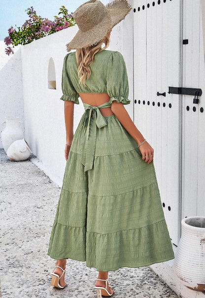 Woman wearing a midi dress with puff sleeves and a tiered skirt, styled with white heels and a straw hat in a Mediterranean setting.