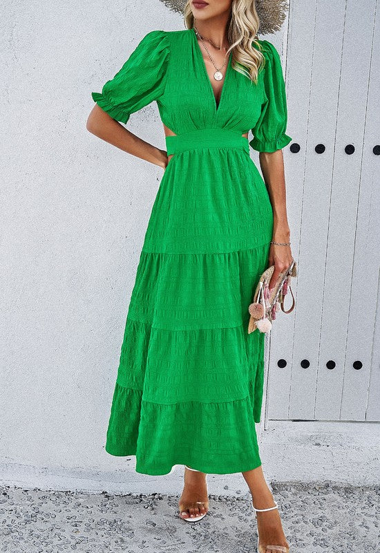 Woman wearing a midi dress with puff sleeves and a tiered skirt, styled with white heels and a straw hat in a Mediterranean setting.