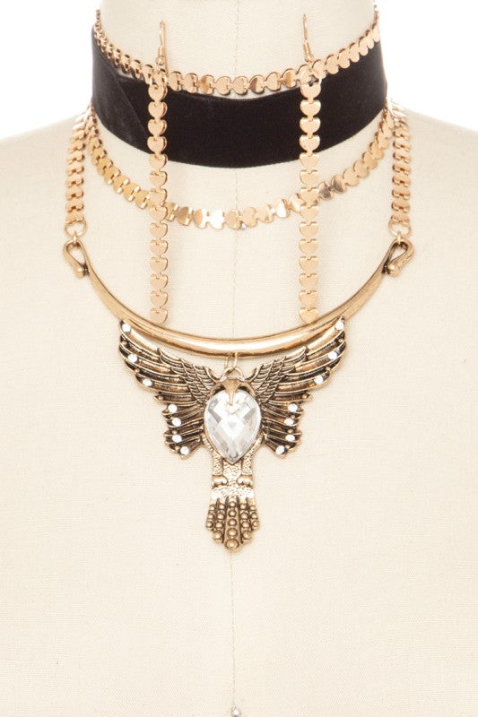 A stylish layered necklace on a mannequin. There's a black velvet choker with gold accents at the top, followed by multiple gold chain layers. The main piece is a gold necklace with a pendant featuring a clear gemstone surrounded by wing-like details and a tassel at the bottom.